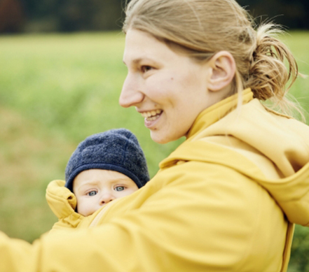 Eine junge Mama mit gelber Tragejacke hält lächelnd ihr Baby an der Brust.