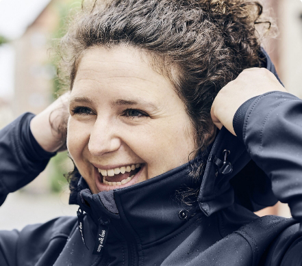 Eine junge Frau stellt sich lachend den Kragen ihrer schwarzen Tragejacke nach oben.