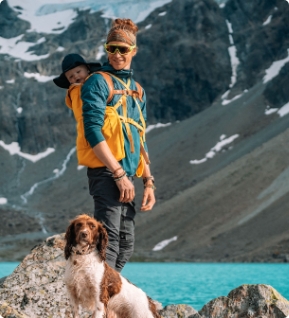Ein junger Papa mit Baby am Rücken in Trage und Hund in den Bergen an einem See.